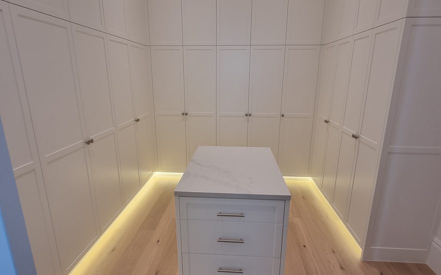 Walk-in wardrobe with white shaker built-in robes, LED toe-kick lighting, and a central stone-topped island