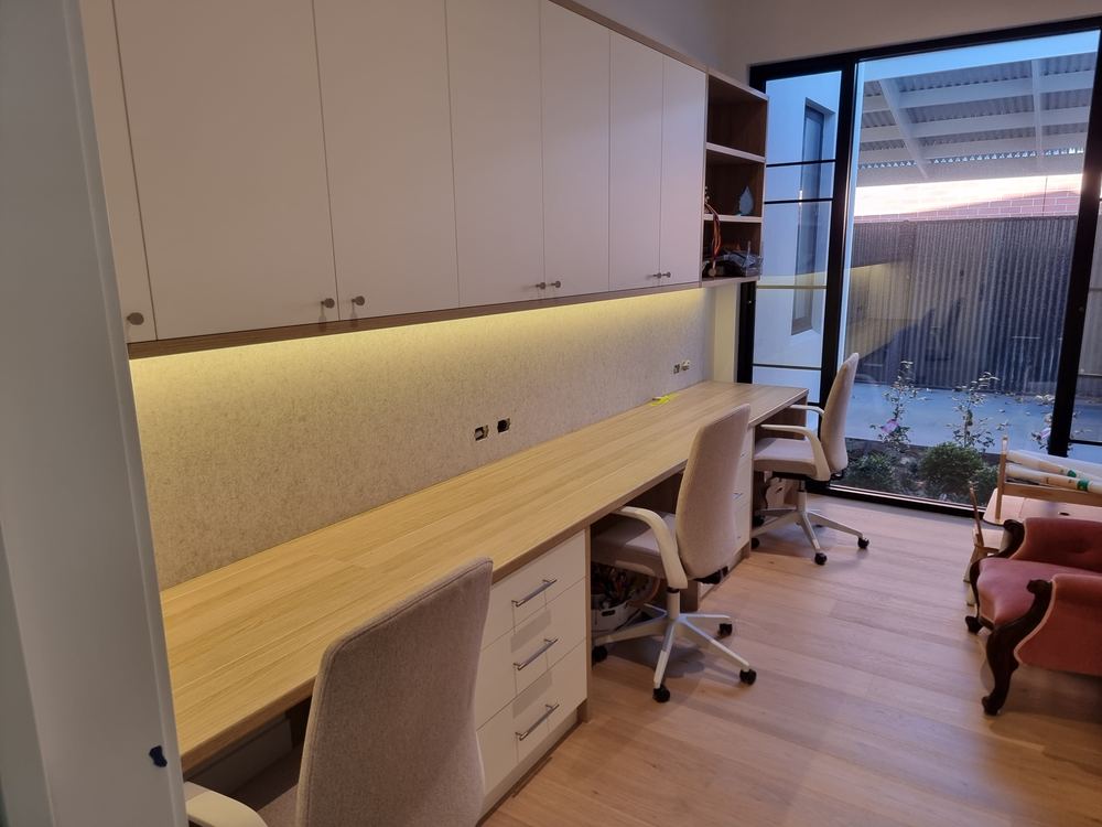Custom home office with a long timber desk, white overhead cabinetry, under-cabinet LED lighting, and drawer storage beside a window