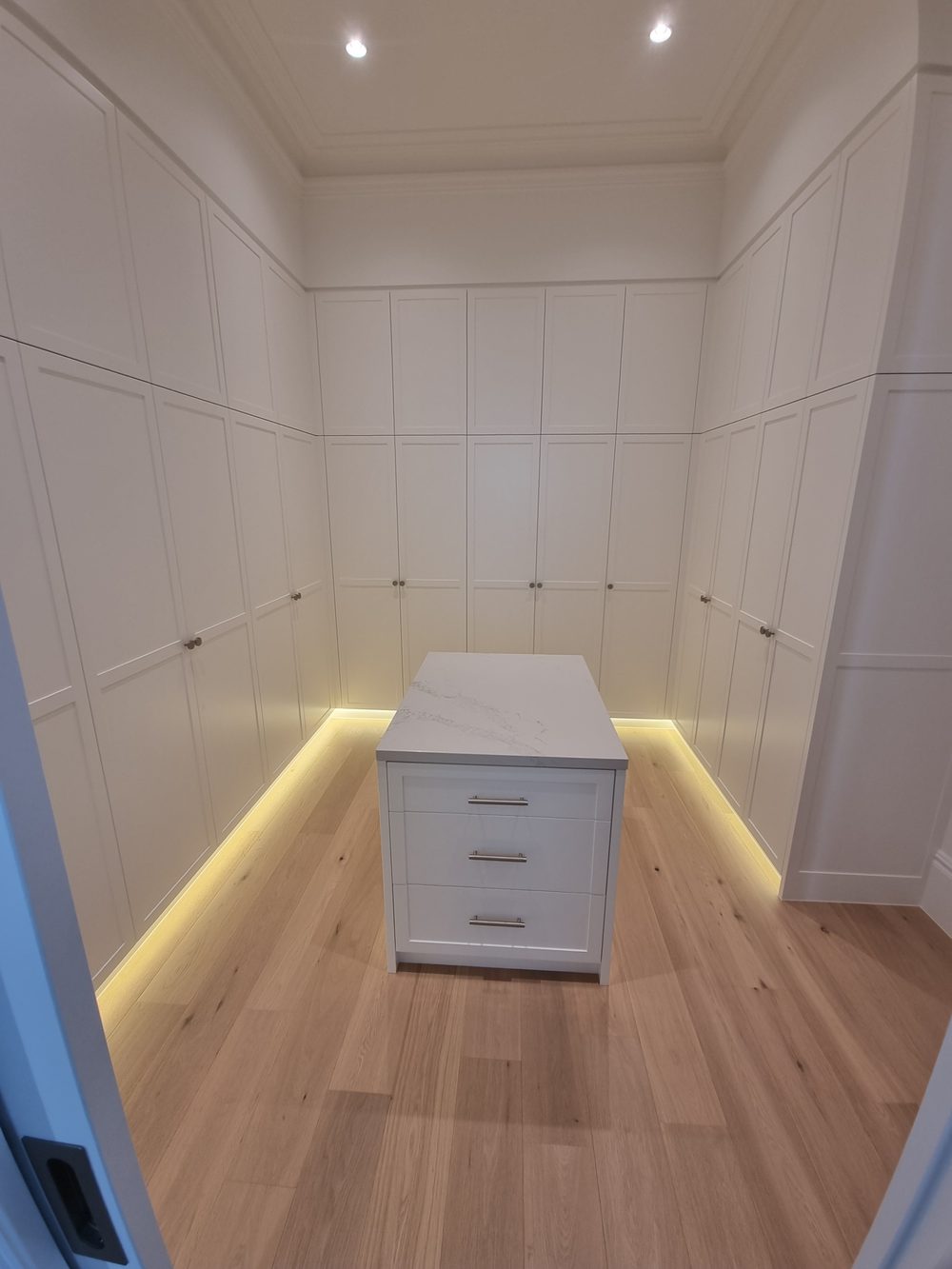 Walk-in wardrobe with white shaker built-in robes, LED toe-kick lighting, and a central island