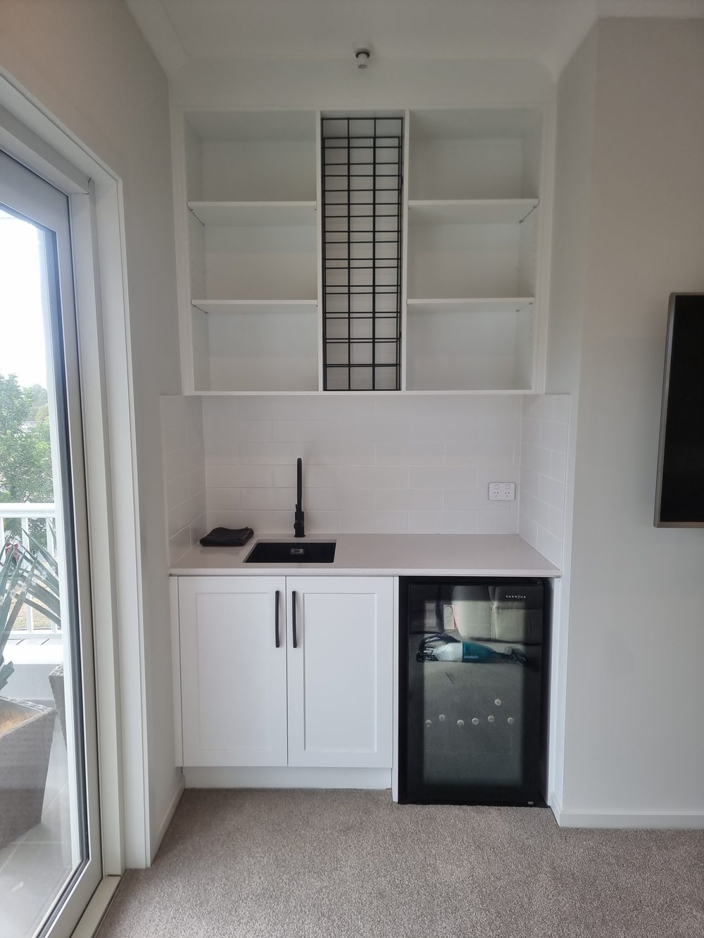 Custom built-in kitchenette with white shaker cabinetry, open shelving, a bar sink, and an integrated bar fridge beside a balcony door