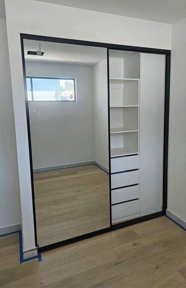 Black-framed sliding mirror wardrobe with fitted internal shelving and drawers beside a window