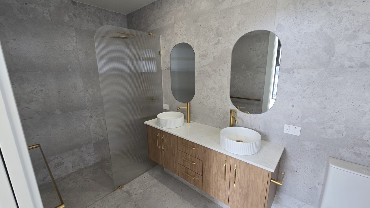 Bathroom with a floating timber double vanity, round fluted basins, brass tapware, and a fluted glass shower screen against stone-look tiles