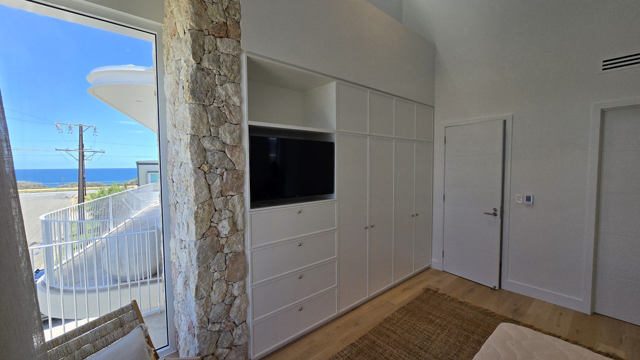 Custom white built-in wall unit with a TV recess, drawers, and full-height wardrobes beside a stone feature wall and an ocean-view balcony