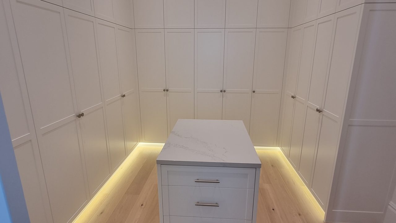 Walk-in wardrobe with white shaker built-in robes, LED toe-kick lighting, and a central stone-topped island